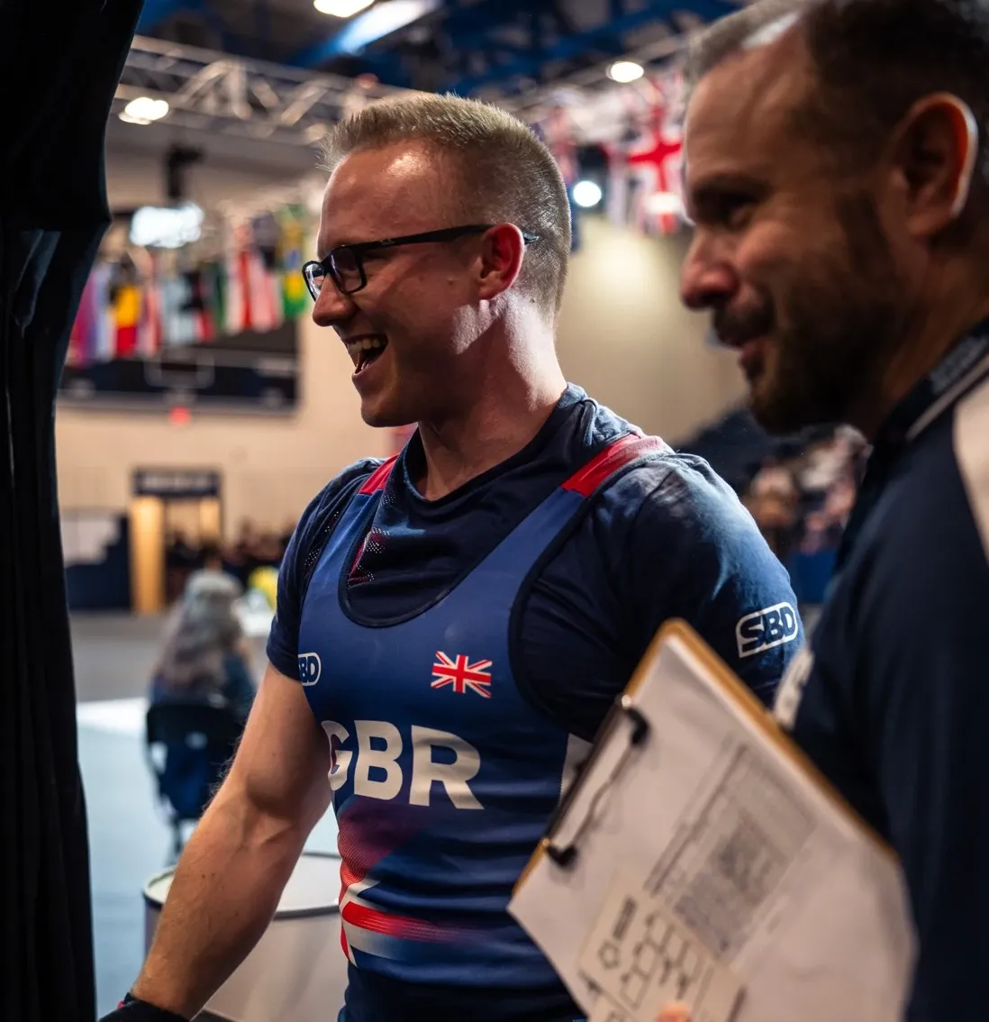 Andrew in the Great Britain singlet, laughing as he walks into the warm-up area after his personal best, next to a team official holding a clipboard